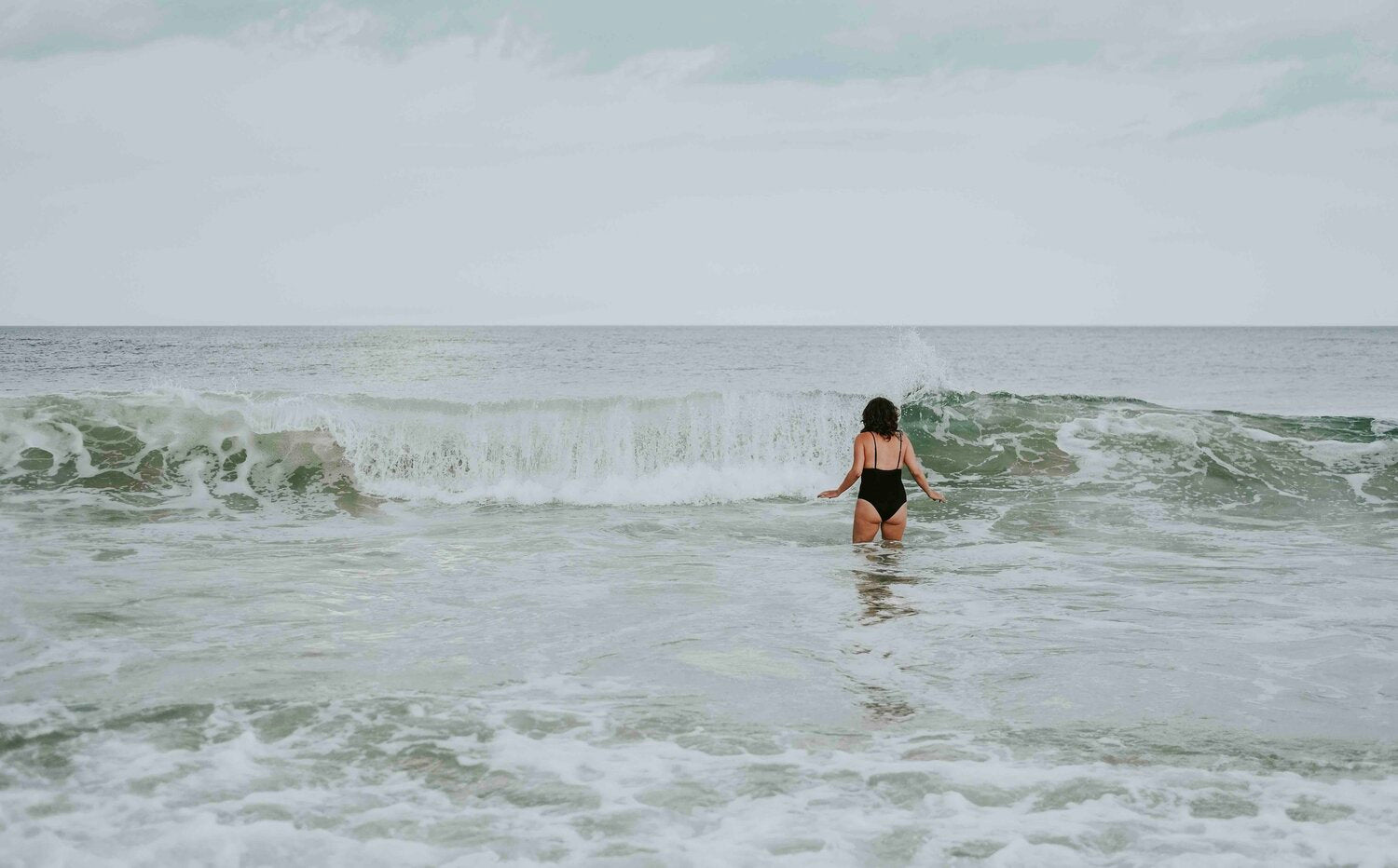 Woman wading into sea waves in a black swimsuit, the salt water and sun exposure behind a post summer skin reset and sun damaged skin repair routine