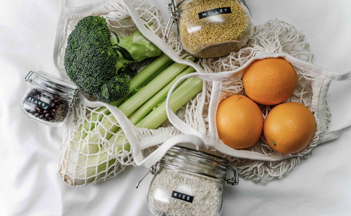 Zero waste flatlay showing a mesh produce bag of fruit and vegetables beside glass storage jars, illustrating circular beauty impact and rescued ingredients
