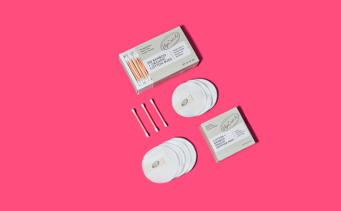 Flatlay of plastic-free bathroom swaps including a safety razor, bamboo cotton buds, reusable makeup pads and beauty accessories on a bright pink background