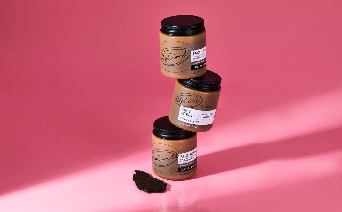 Three UpCircle glass skincare jars stacked on a pink background with loose upcycled coffee grounds, an example of sustainable skincare packaging in glass and aluminium