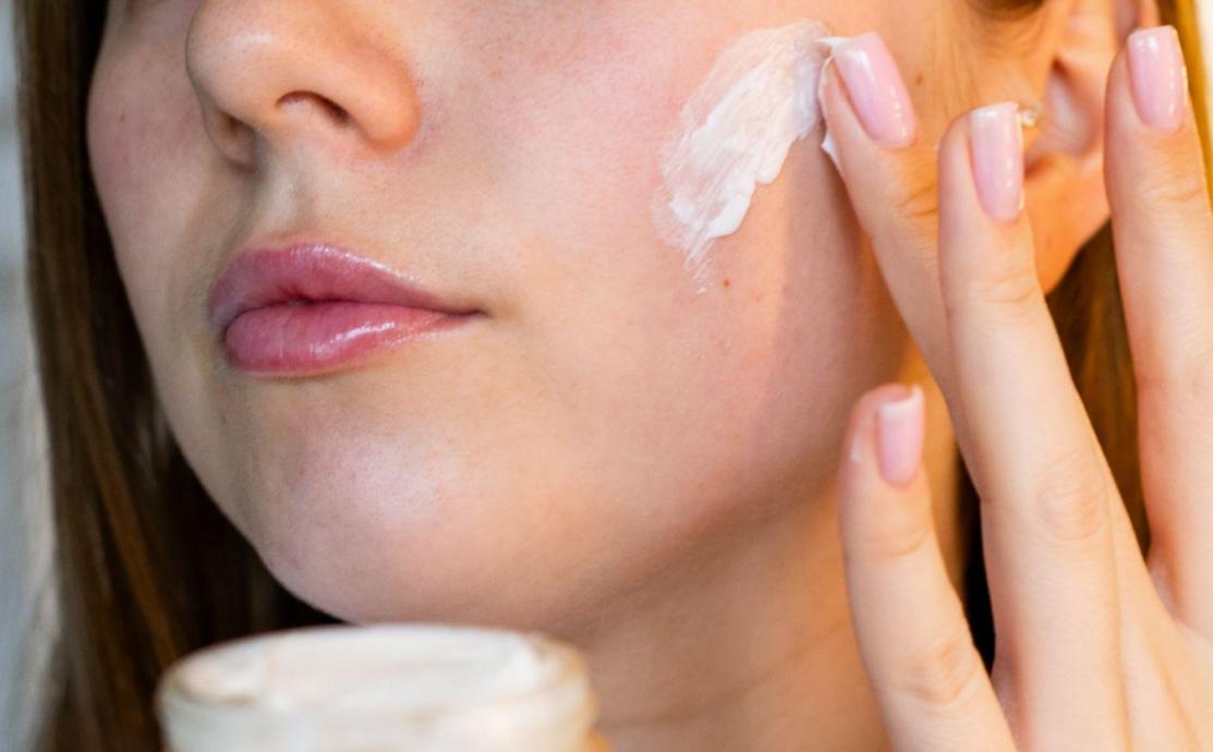 Woman applying moisturiser to her cheek as part of a skincare routine for over 50, with a jar of natural mature skin cream in the foreground